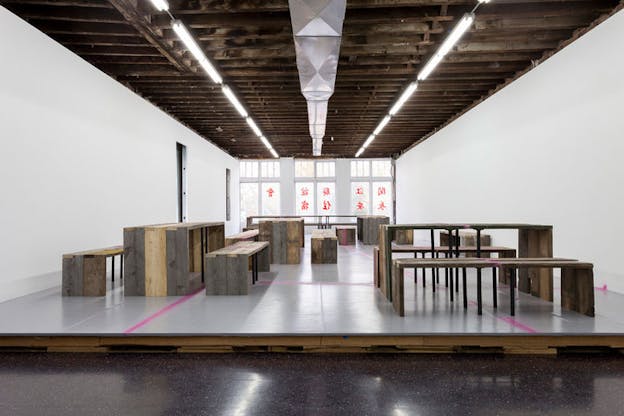 An installation image of five sets of tables and benches constructed out of thick plywood panels hued medium brown or grey. These sets each have different orientations and rest on a grey tiled floor which is elevated by wooden planks off of a black and white speckled floor. A bubblegum pink line runs across the floor and over some furniture. The ceiling is exposed wood planks and in the background, there are floor to ceiling windows printed with red Chinese characters. 