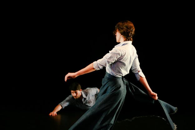 A performance still of two performers in a darkened space. The performer in the foreground has braided hair and wears a button down shirt and a long skirt. They face away from the audience and hold out their skirt behind them. The second performer wears a button down shirt and crawls on the black floor with one arm outstretched in front of them and the other behind. 