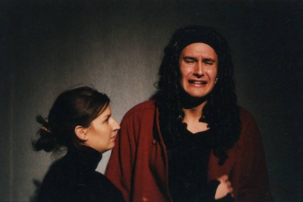 Two performers are in front of a grey background. The person on the right has a distressed expression on their face. They have long brown hair and wear a red coat. The person on the left wears a black turtleneck and looks to the distressed person and reaches toward them.