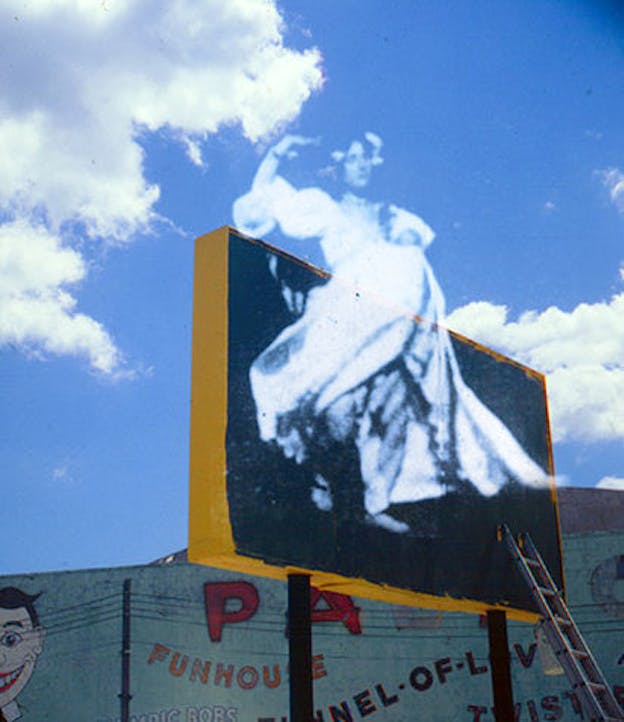 Picture of a billboard and blue cloudy sky. A transparent film still projection of a silhouette is stamped on the front.