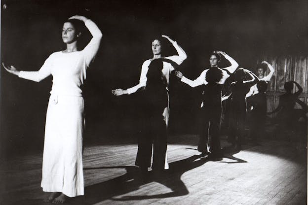 Black and white still of five aligned performers closing their eyes and raising their arms in a crescent shape, each performer's body shadowed by the performer in front of them.