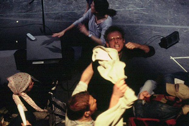An overhead photograph of a performance taking place on a black stage. People appear flustered and are crowded in the foreground of the image. One person holds a fabric above their head defiantly. Another person looks up towards the camera and has a raised fist. 