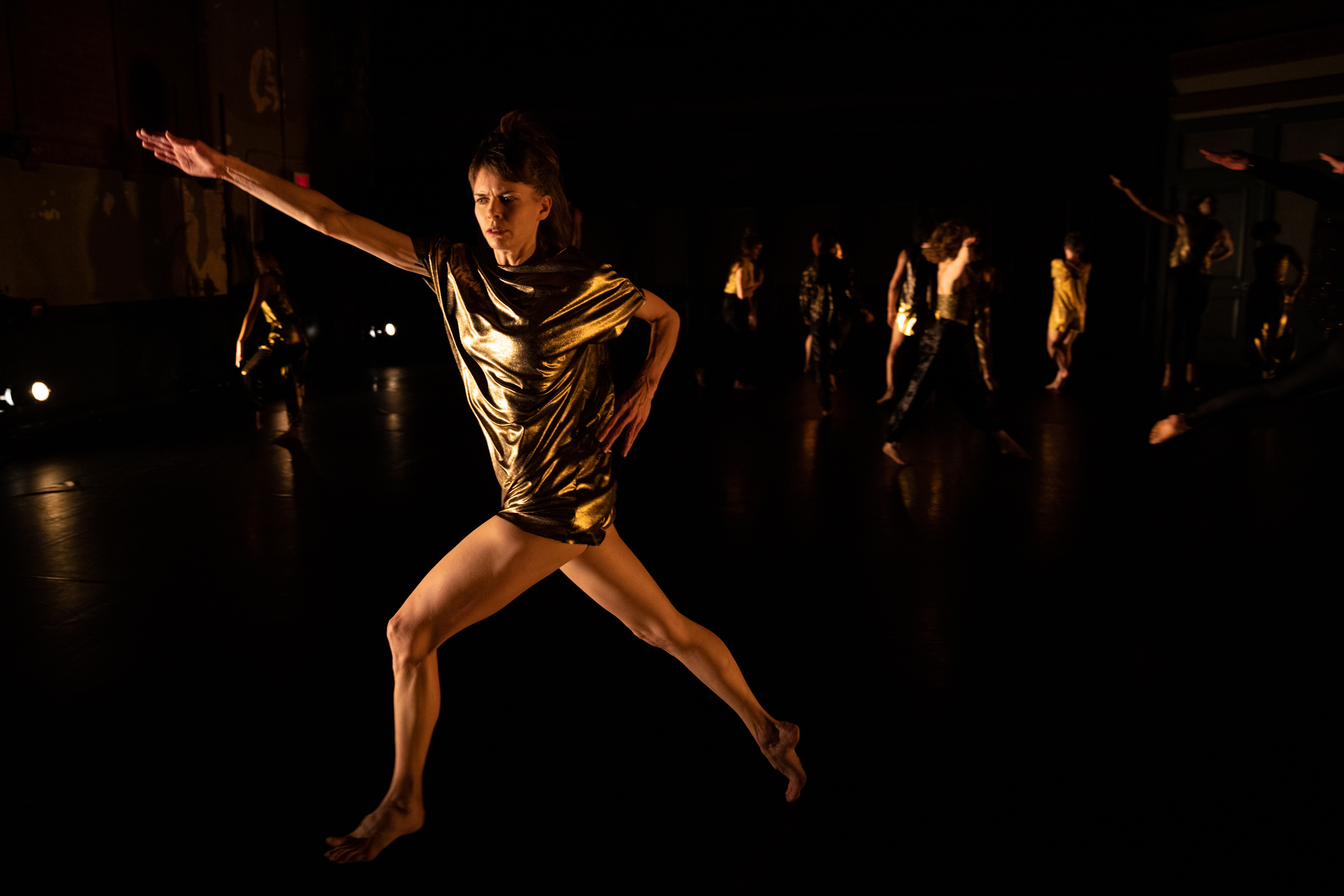 Joanna Kotze is centered as she performs on a dark stage with minimal yellow lighting, some dancers are visible in the far background behind her. She looks towards the left with concentration as she lunges forward on her left leg, hips pointing to the left. Her torso twists as her shoulders angle towards the camera. Her left arm is bent as her hand rests on her left hip, and her right arm is extended and reaches out and up to the left where she lunges, hand outstretched with her palm facing down. She wears a gold metallic shortsleeved tunic.