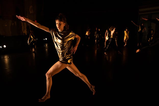 Joanna Kotze is centered as she performs on a dark stage with minimal yellow lighting, some dancers are visible in the far background behind her. She looks towards the left with concentration as she lunges forward on her left leg, hips pointing to the left. Her torso twists as her shoulders angle towards the camera. Her left arm is bent as her hand rests on her left hip, and her right arm is extended and reaches out and up to the left where she lunges, hand outstretched with her palm facing down. She wears a gold metallic shortsleeved tunic.