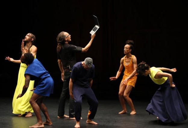 Performers dressed in colors of blue, yellow and orange move on stage in various poses and surround a figure dressed in black. The performer in the middle 
