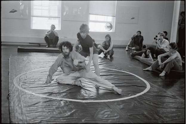 Black and white photograph of two people moving inside a white circle drawn on the floor while others seated down around it watch them.