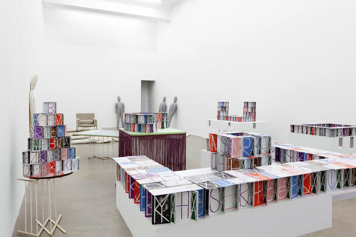 Installation view of a white room filled with tables hosting rows of multi-colored boxes displaying juxtaposed letters and shapes. At the far-end of the room, gray blow-up dummies are rested against the walls