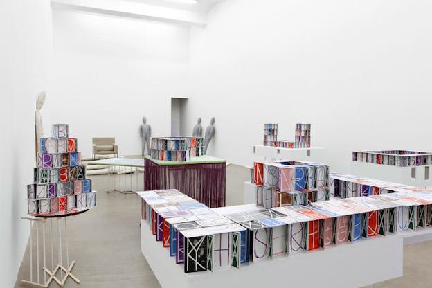 Installation view of a white room filled with tables hosting rows of multi-colored boxes displaying juxtaposed letters and shapes. At the far-end of the room, gray blow-up dummies are rested against the walls