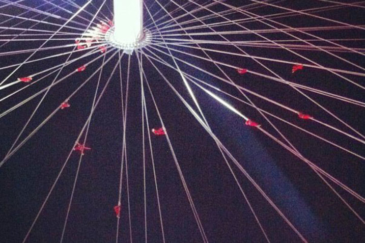 Multiple cables that support performers clad in black meet in the middle of a white column.