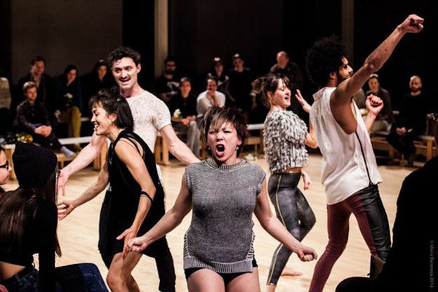 Dancers on a floor-level stage surrounded on all sides by a benched audience dance emphatically: one raises their fist in the air, and one facing the camera opens their mouth like a roar, bending their knees and arms.