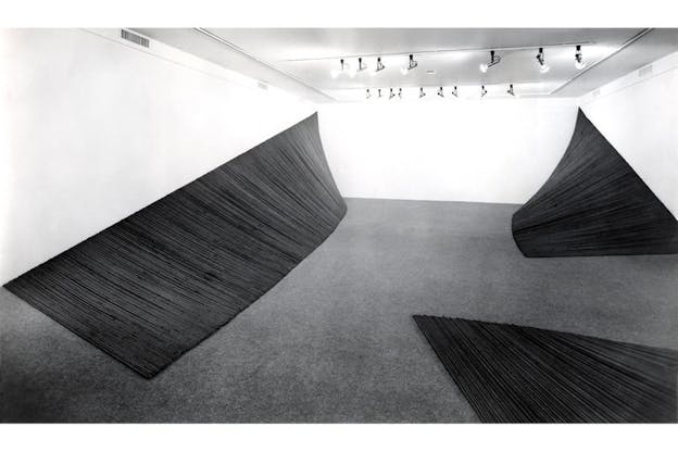 Installation view of three large black sculptures curved like paper like in a blank room with gray floors. 