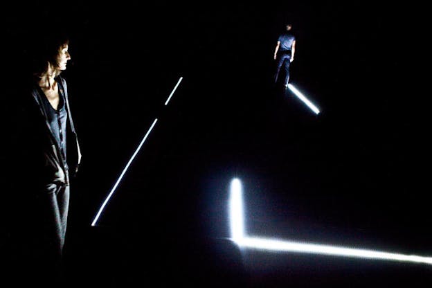 Two performers stand in a dark room, lit only by a few lines of white light. In the foreground, a performer stands on the left, wearing street clothes with their hands in their pockets, looking downwards. In the background, a performer stands with his back to the camera. 