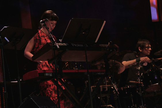 Low-angle shot of Mazzoli in a red and black printed dress performing on keyboard beside Glenn Kotche on drums. 