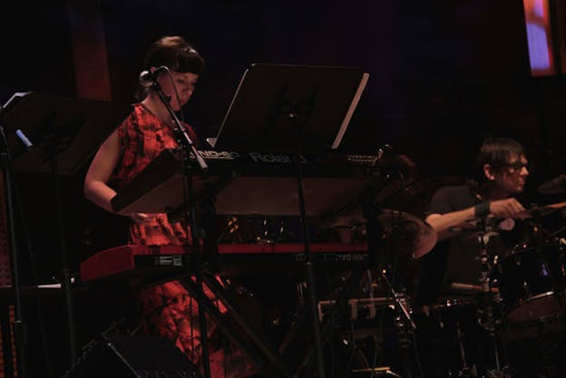 Low-angle shot of Mazzoli in a red and black printed dress performing on keyboard beside Glenn Kotche on drums. 