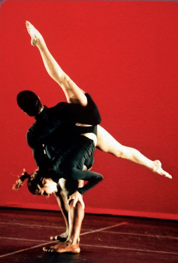 Two performers wearing all black are situated against a bright red background. One performer has their back turned to the camera while the other performer holds onto that first performer's leg to support their entire body as it is held upside down. 