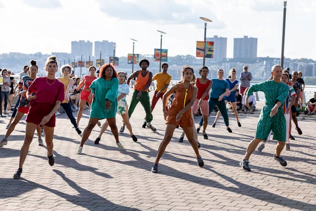 A large group of dancers wearing an array of colorful clothing perform facing the camera. They are outdoors in the sun on a pier with a city skyline visible in the background. All executing the same movement in sync, their legs are straight as they stand upright with their feet apart. Their bodies lean slightly to their left, their left arms are reaching diagonally across their bodies with straight hands while their right arms are curved by their sides with ther elbows pointed out and behind them. 