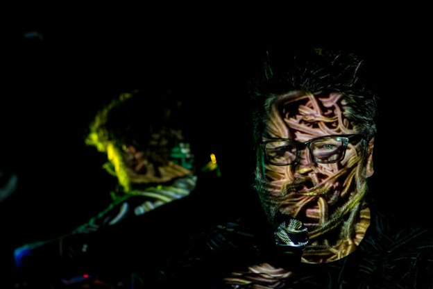 Close up of a performer in a black-lit room, their face projected with fleshy pink and varying green intestine-shaped imagery. 