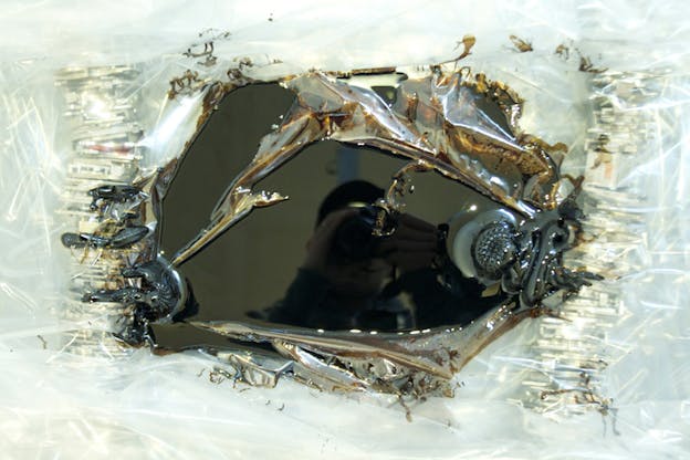 A white glass surface centers around a black glass one with hues of brown and elevated patterns on the outline. The reflection of a person with a camera covering their face can be seen on the black surface.