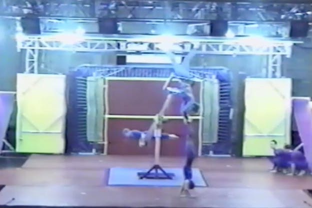 A performer is supported mid-air by a mechanism that surrounds his waist. His body is in a horizontal angle while another gymnast grabs his legs.