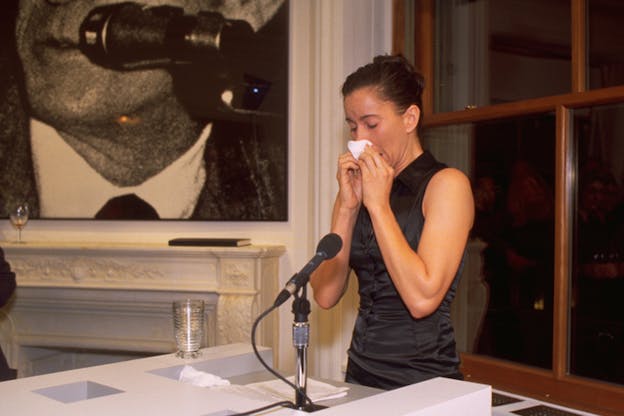 A figure dressed in a black satin dress with an updo standing behind a desk full of papers and a microphone, holding a tissue in front of their nose.  