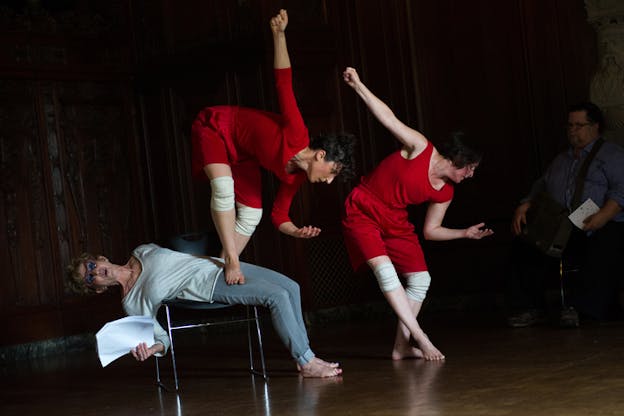 Two performers dressed in red crouching similarly with one hand angled and the other extended upwards. One of the performers balances themselves on top of a third figure laying sideways on a chair.