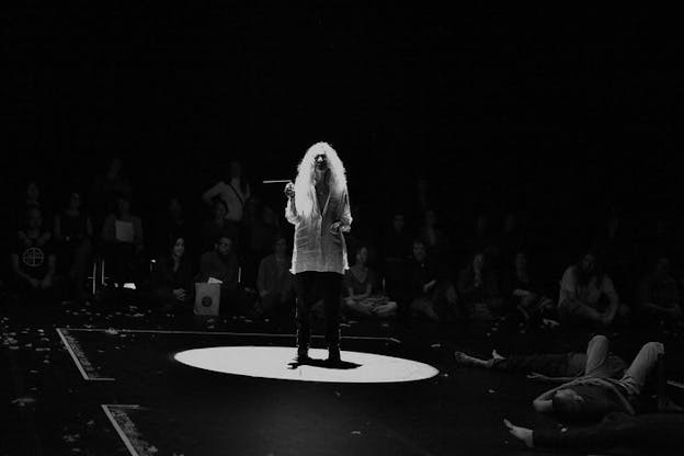 Black and white photograph of a figure with long light curly hair, a light long button up and dark pants, standing in the middle of a stage illuminated by a circle. 