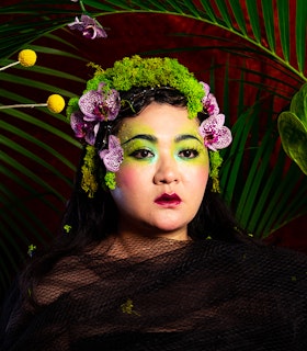 A portrait of Du Yun surrounded by green palm tree leaves. Dressed in black tulle material, the artist wears a crown of green moss and magenta orchids. The make-up surrounding her eyes is a heavy neon green color.
