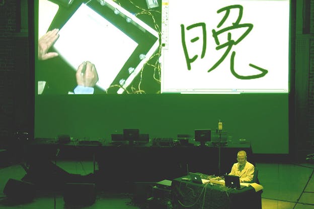 A green-tinted photograph of a performance with Yasunao Tone behind a black desk covered in laptops and wires. Behind him, there is a longer desk covered in computers and on the wall, there is a split projection with his hands drawing something on the left and an alphabetical character on the right.