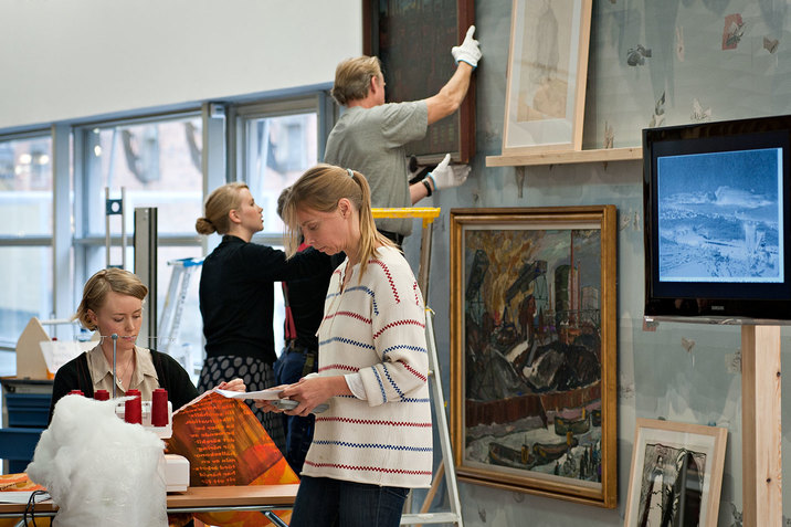 Backstrom glances down at a sheet of paper and stands beside a person sewing an orange banner while behind them, two persons hang framed paintings on a wall. 