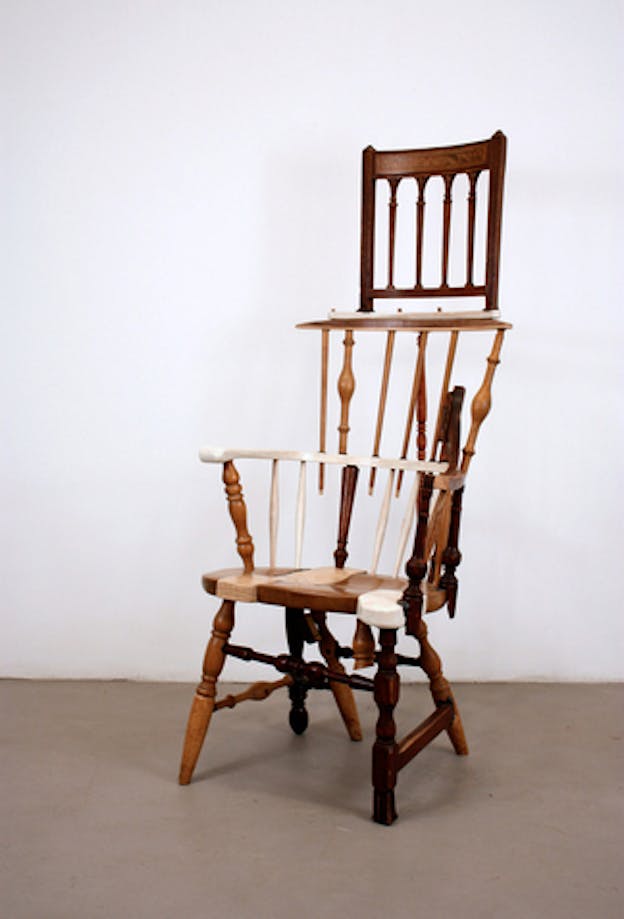 A sculpture made of a wooden chair with the backs of two other wooden chairs stacked on top of it. The sculpture is in an otherwise bare room with a white wall and grey floor. 