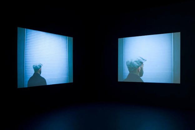 Two side-by-side slanted blue-hued images suspended in black space, one close-up and one distant, of a person with their back turned wearing a pageboy cap and facing a white grated wall. 