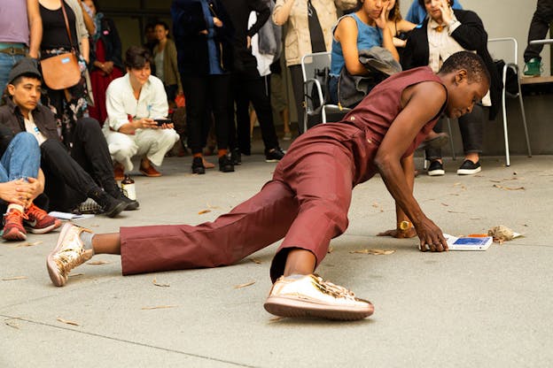 A performer clad in burgundy supports their upper body with their hands while their bottom half lies on the floor. They look down towards a book with a white and blue cover. an audience standing and sitting down on the floor surrounds them.