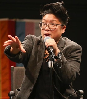 Portrait of Kay Ulanday Barrett standing and speaking into a microphone. They wear a grey blazer, round glasses, and have short black hair. A transgender flag and a queer pride flag hang from the wall behind them.