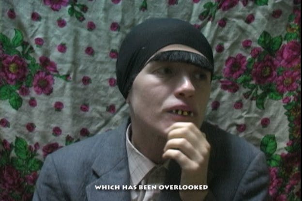 A video still of Ben Tor in front of a floral background, wearing a black cap, fake bushy eyebrows, a fake set of teeth (with some missing) and a grey suit jacket and pink plaid shirt. She holds her chin in her hand and looks into the distance. A video still of Ben Tor in front of a white wall, wearing a disorganized brown wig with scattered bits of hair on her face, a fake nose, and a grey suit jacket with a blue shirt. She hunches her shoulders and smiles at the camera. 