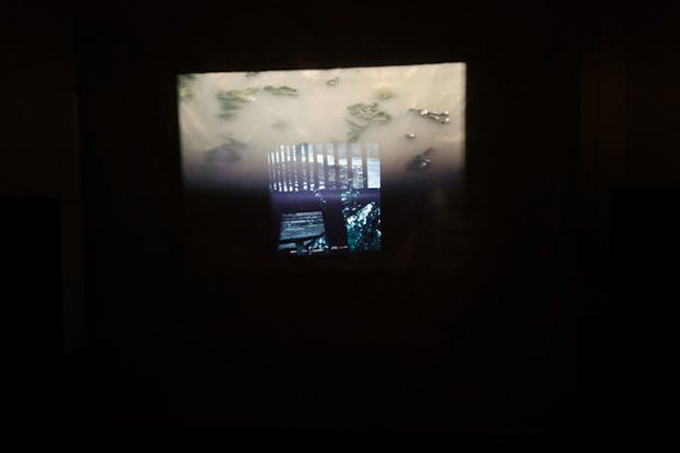 Rectangular image of the underside of a wooden balcony divided by a t-shaped metal beam from a transparent, juxtaposed image of black railings superimposed on a rectangular image of muddy water and floating leaves, suspended in black space. 