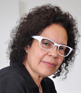 Portrait of Candida Alvarez, with medium-length dark curly hair, wearing white-framed glasses and a black collared shirt.
