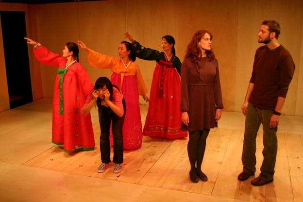 Three people dressed in traditional Korean wear stretch their right arms out at a diagonal and gaze beyond them. One person in modern wear stands in front of them, grimacing forward in a hunched position. To the right of them, two people dressed in brown-toned clothes stare at each other. The set has wood floors and walls and an open doorway to darkness in the back left corner.