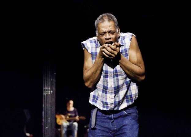 Houston-Jones wearing a flannel and blue jeans making a coughing motion into two clasped palms on a dark stage where in the background a guitarist is blurred. 
