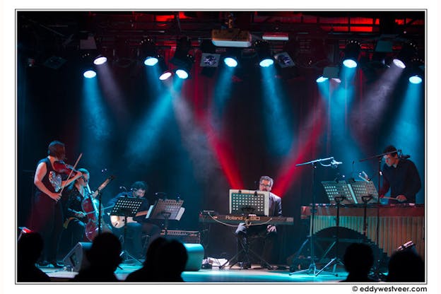 A band composed of violin, cello, bass, keyboard, and percussion performs on stage under red and blue spotlights.