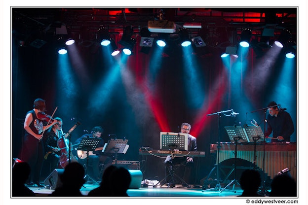 A band composed of violin, cello, bass, keyboard, and percussion performs on stage under red and blue spotlights.