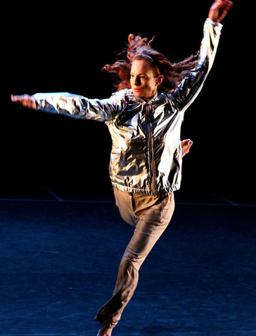 Person with loose red hair wearing a reflective silver jacket and brown pants leaps with one leg bent backwards and one extended forwards, raising their arms in an L-shape and looking outwards.