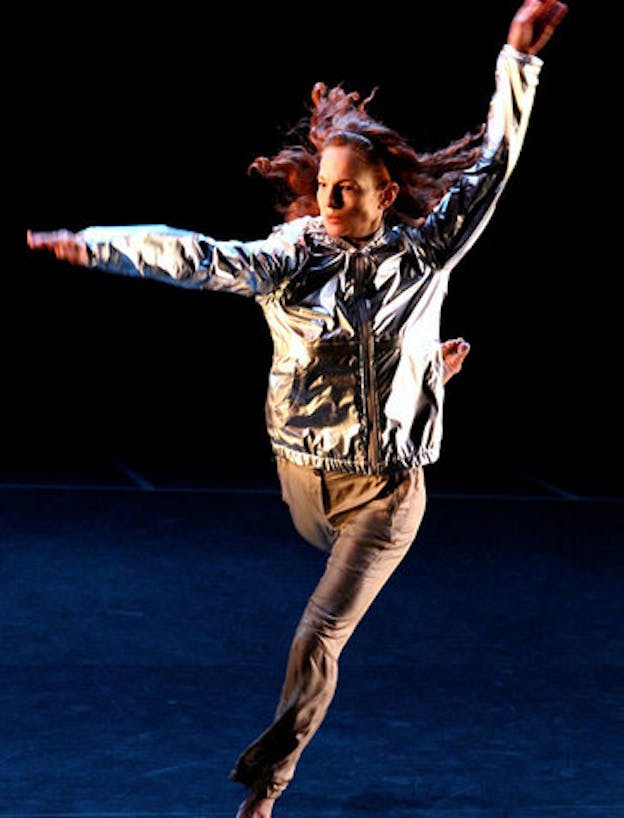 Person with loose red hair wearing a reflective silver jacket and brown pants leaps with one leg bent backwards and one extended forwards, raising their arms in an L-shape and looking outwards.
