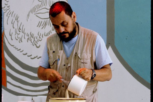 A person with red hair wearing a tan vest and blue tee-shirt concentrates on whisking something in a white bowl as they stand in front of a mural of round blue shapes and a black and white sketch of a man's beard.