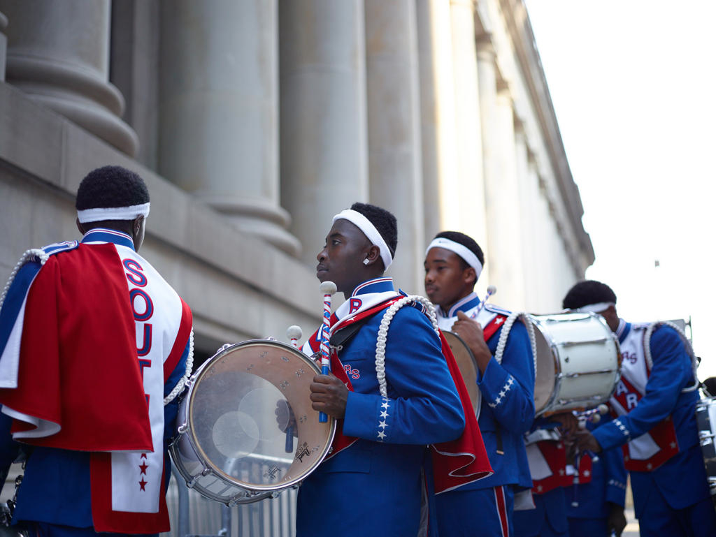 Young people in red, white, and blue marching band uniforms, carrying marching drums, and filing in a line in front of a stone columned building.