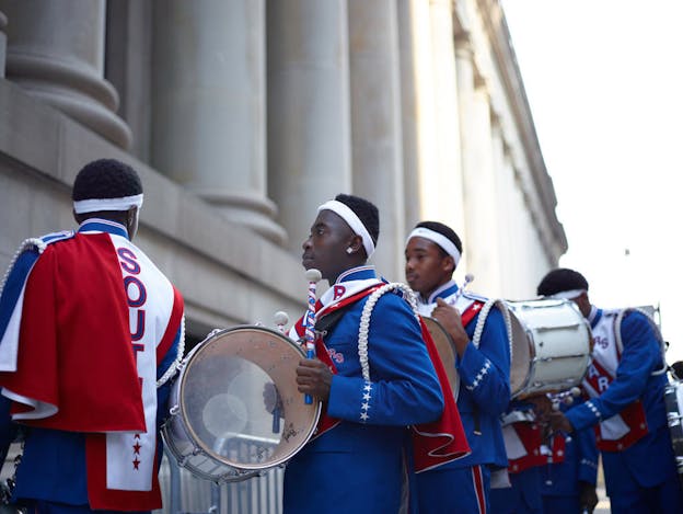 Young people in red, white, and blue marching band uniforms, carrying marching drums, and filing in a line in front of a stone columned building.