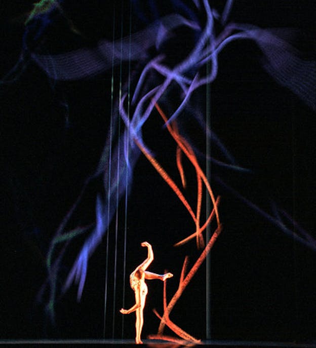 One performers dances in the lower middle of a tall dark stage. They stand on only one leg and bend towards the right with the rest of their body. In the background, there is a projection of a red and purple sketch of a human figure lunging towards the right.