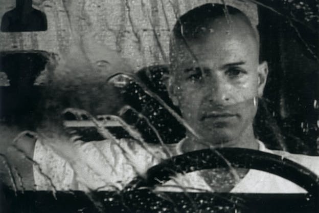 Black and white image of a person standing behind a car wheel in a black and white photograph. The glass in the front has trails of rain going down as the person looks in the distance with a grim expression.
