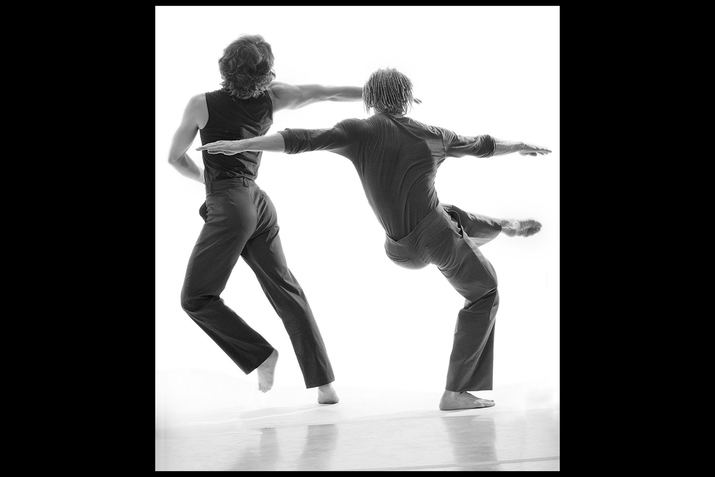 A black and white performance still of two dancers in a white space. The dancers wear all dark clothes and face away from the camera. The dancer on the left bends slightly backwards and leans to the left, lifting their left foot. The performer on the right bends on the right foot and lifts their right foot upwards by their waist.