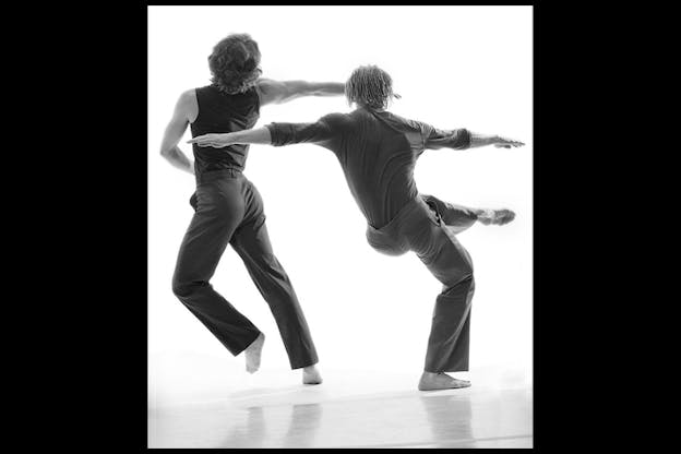 A black and white performance still of two dancers in a white space. The dancers wear all dark clothes and face away from the camera. The dancer on the left bends slightly backwards and leans to the left, lifting their left foot. The performer on the right bends on the right foot and lifts their right foot upwards by their waist.