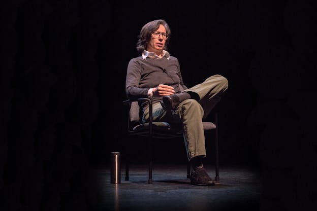 A figure dressed in beige pants and a gray sweater with glasses and graying mid-length hair, sits on a chair with one leg resting on the other.